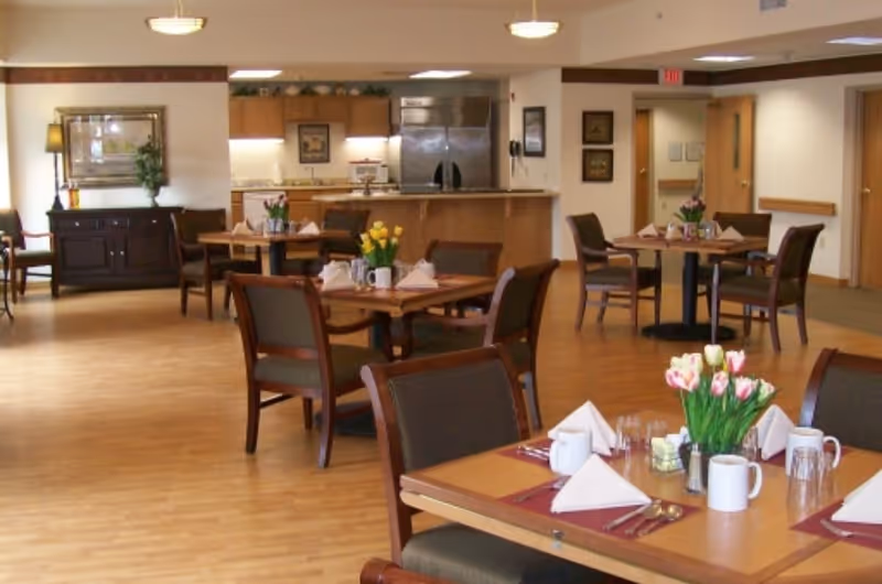 A dining area in a senior living facility with several wooden tables and chairs. Each table is set with white napkins, mugs, glasses, and silverware. There are flower arrangements on the tables. In the background, there is a kitchen area with a stainless steel refrigerator and wooden cabinets. The room has wooden flooring and soft lighting.