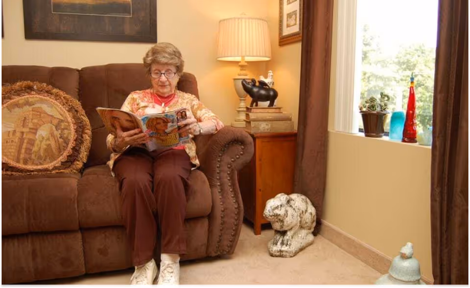An elderly woman sitting on a brown couch reading a magazine in a cozy living room. The room features a side table with a lamp and decorative items, a window with brown curtains, and various decorative objects on the floor and windowsill.