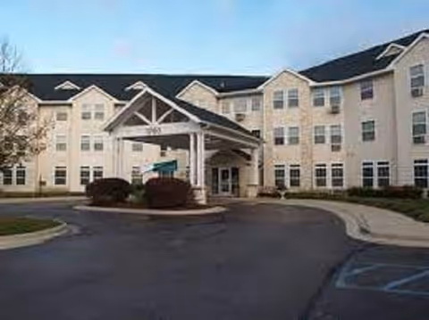 Exterior view of a large, multi-story senior living facility building with beige siding and multiple windows. The entrance features a covered drop-off area with a peaked roof supported by columns. There are bushes and a paved driveway in front of the building under a clear sky.