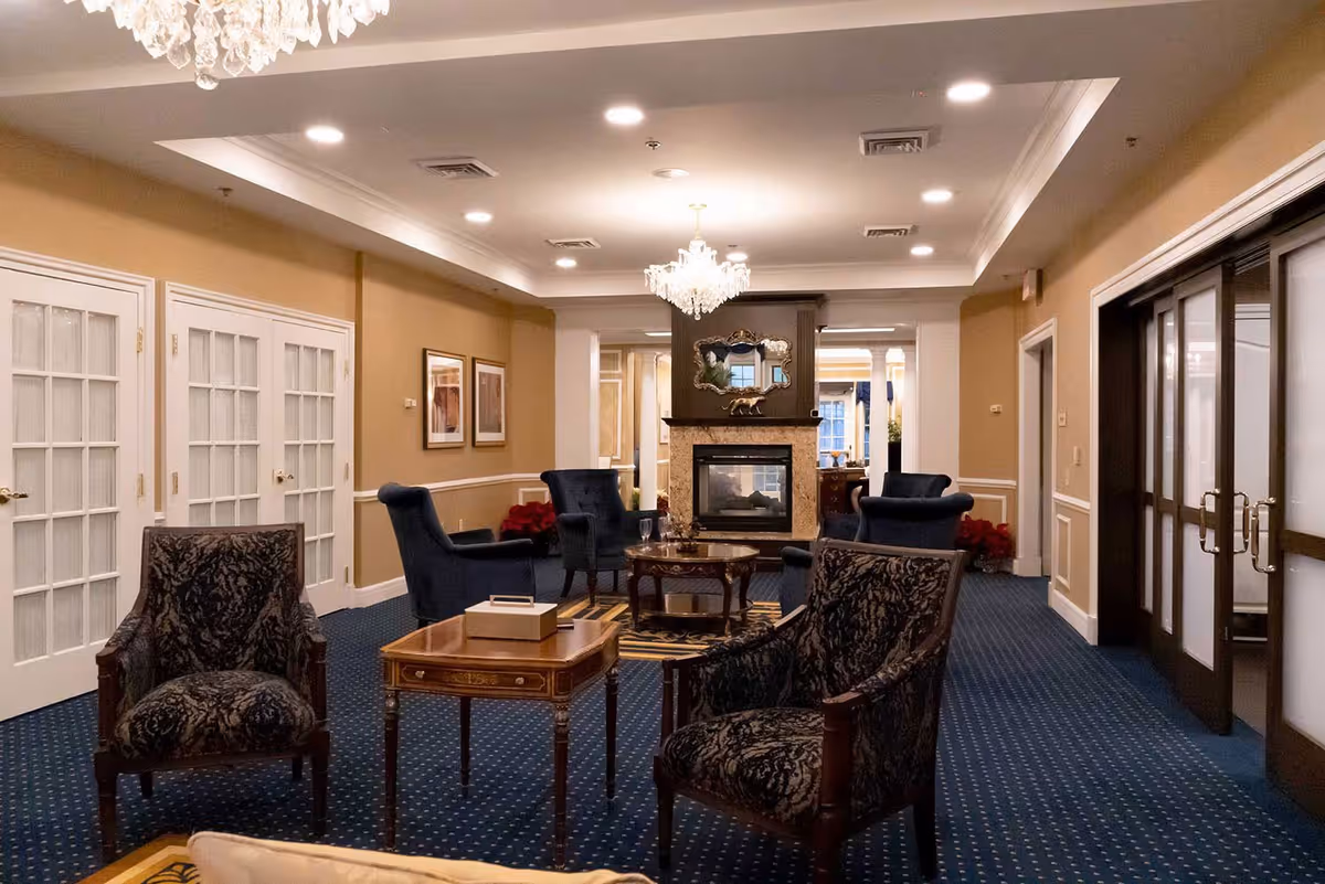A cozy and elegant common area featuring a blue carpet with a dotted pattern, several upholstered armchairs in dark colors, a wooden coffee table with a tissue box, and a fireplace with a decorative mirror above it. The room has beige walls with white trim, recessed ceiling lights, and a chandelier hanging from the ceiling. French doors and large sliding doors are visible on the sides.