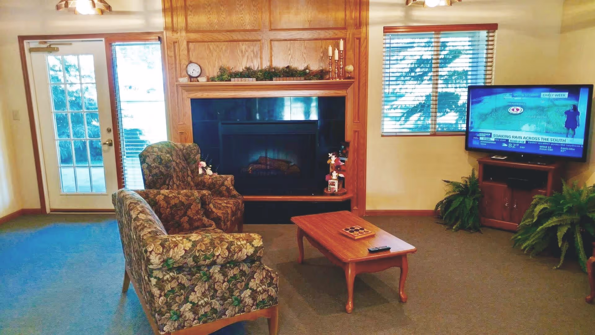 A cozy living room with floral patterned armchairs and a matching loveseat arranged around a wooden coffee table. There is a fireplace with a wooden mantel decorated with greenery and candles. A flat-screen TV is mounted on a wooden stand to the right, showing a weather forecast. The room has a window with wooden blinds and a glass door leading outside. Two green potted plants are placed near the TV stand.