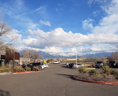 A parking lot with cars and landscaped islands in front of a building, set against snow-capped mountains and a blue sky with clouds.