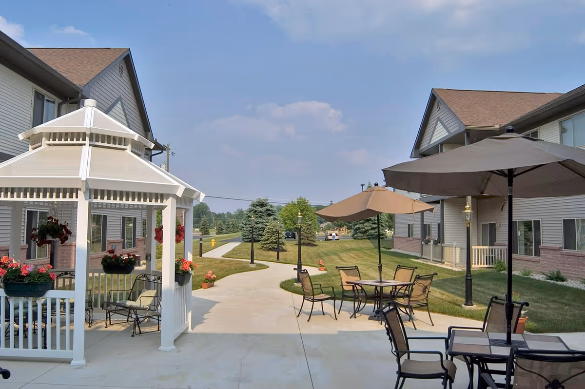 Outdoor patio area at Primrose Retirement Community of Casper featuring a white gazebo with hanging flower baskets, several tables with umbrellas, chairs, and a paved walkway surrounded by grass and trees under a partly cloudy sky.