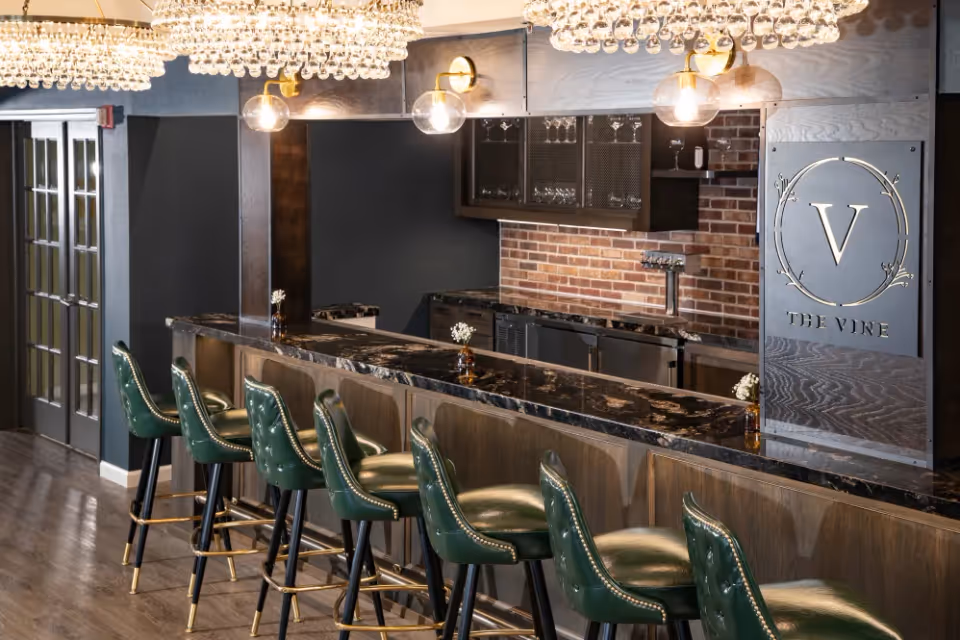 Interior view of a stylish bar area with a long marble countertop and six green leather bar stools with brass nailhead trim. Above the bar are two large chandeliers with hanging crystal balls and two wall-mounted globe light fixtures. Behind the bar is a brick backsplash with shelves holding glassware and a metal sign that reads 'V THE VINE'.