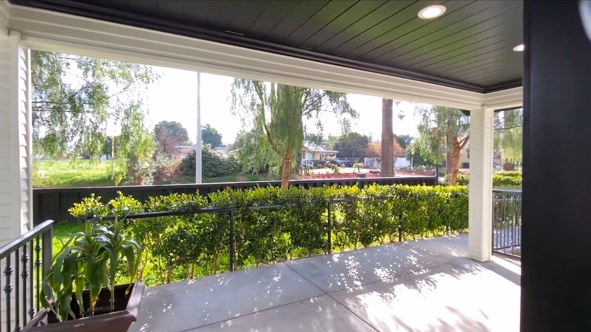 Covered outdoor patio area with a black ceiling and recessed lighting, overlooking a green hedge and trees with a view of neighboring houses in the background.