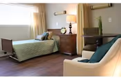 Sunlit bedroom with a single bed, wooden nightstand and lamp, an armchair and small table on wood flooring.