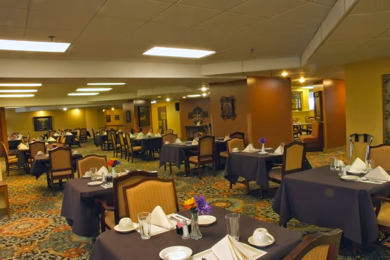 A spacious dining room with multiple tables covered in dark tablecloths, each set with white napkins, plates, glasses, and small flower arrangements. The room has patterned carpet flooring, warm yellow and brown walls, and ceiling lights providing bright illumination.