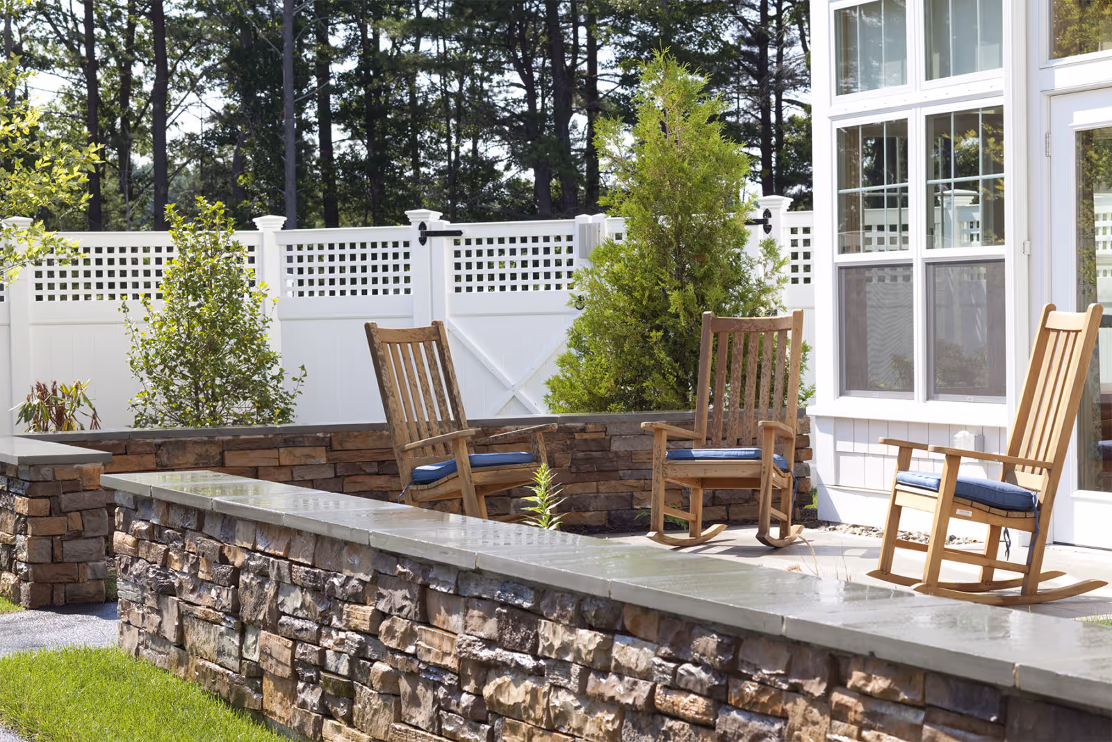 Sunny outdoor patio with wooden rocking chairs on a stone terrace next to a white fence and building windows.