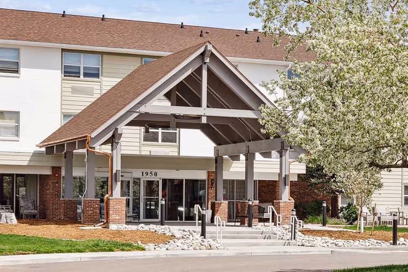 Entrance of a senior living facility with a covered porch supported by wooden beams and brick pillars. The building has a beige and white exterior with multiple windows and a brown shingled roof. There is a tree with white blossoms on the right side and a paved driveway in front.