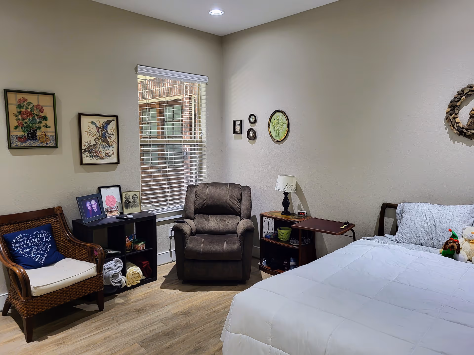 A cozy bedroom with a bed, recliner, wicker chair, side tables, framed wall art and a window with blinds.