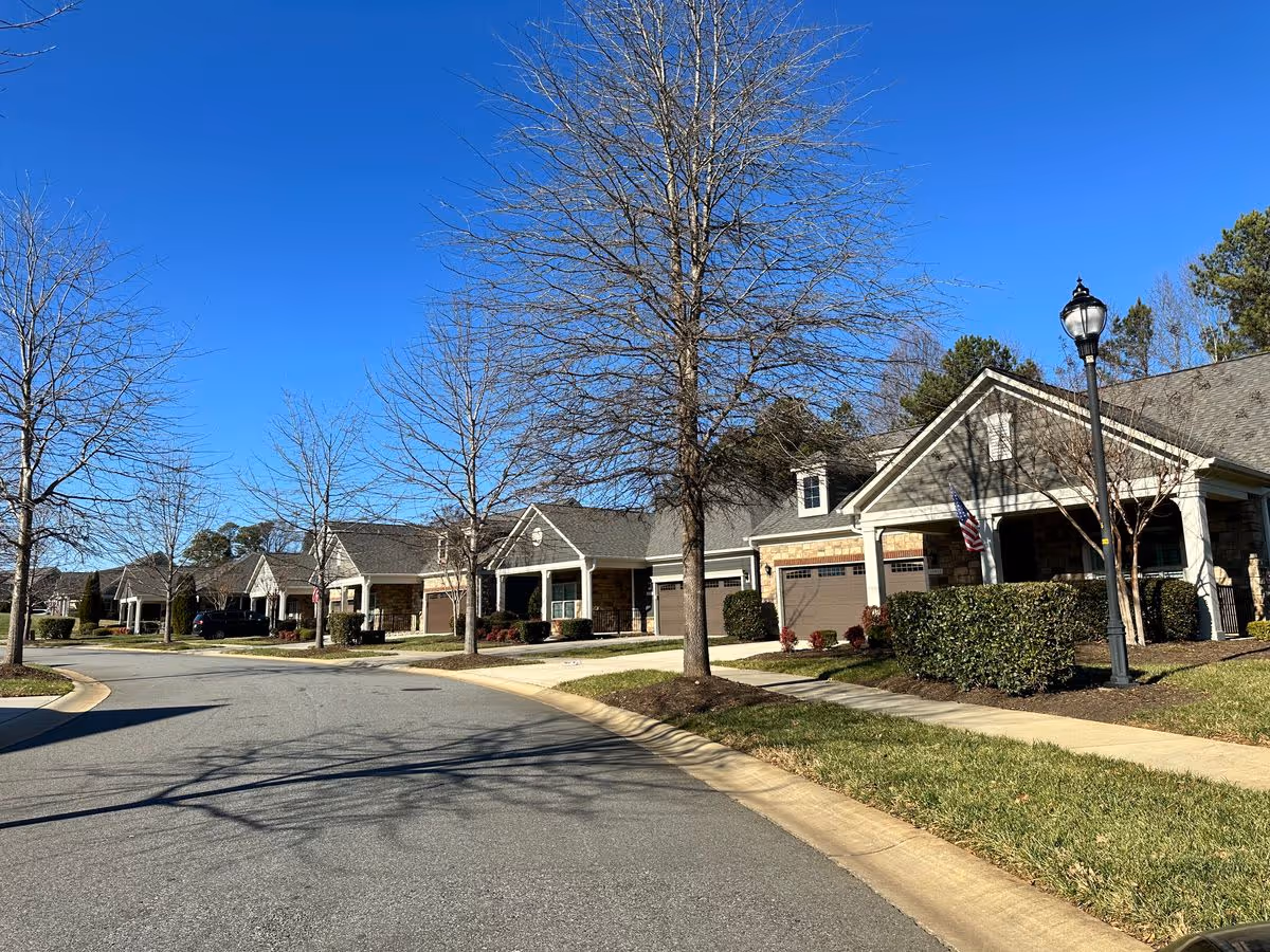 Street view of single-story townhouse-style residences with garages, leafless trees, and a lamppost under a clear blue sky.