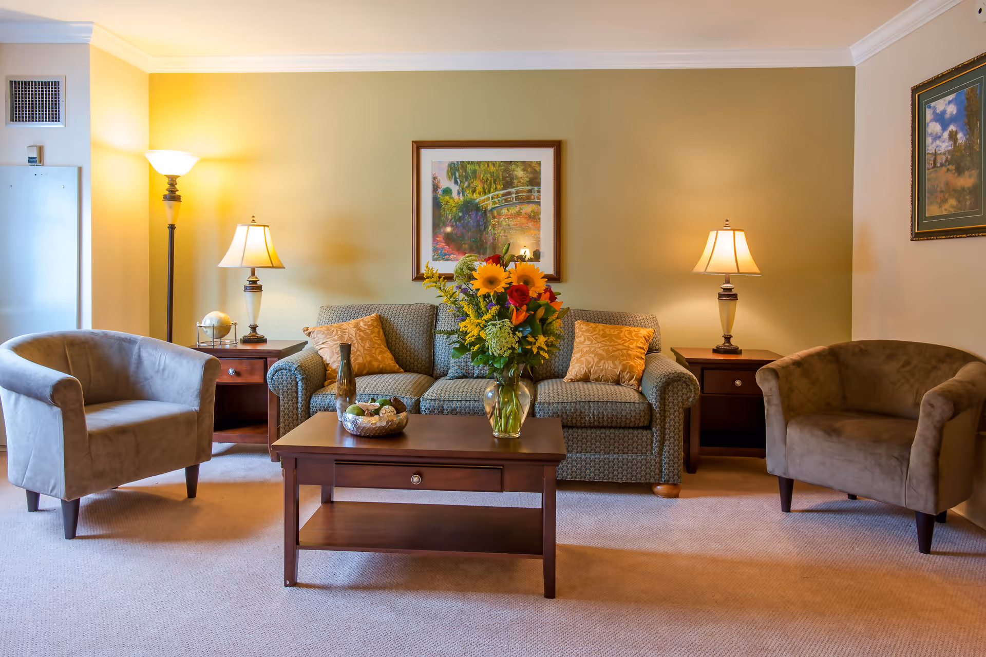 A cozy living room with a patterned sofa adorned with two decorative pillows, flanked by two matching side tables each holding a lamp. Two beige armchairs are positioned on either side of the sofa. A wooden coffee table in front of the sofa holds a vase with a colorful bouquet of flowers and a decorative bowl. The walls are painted light yellow and decorated with framed artwork. The room is softly lit by the lamps and a floor lamp in the corner.