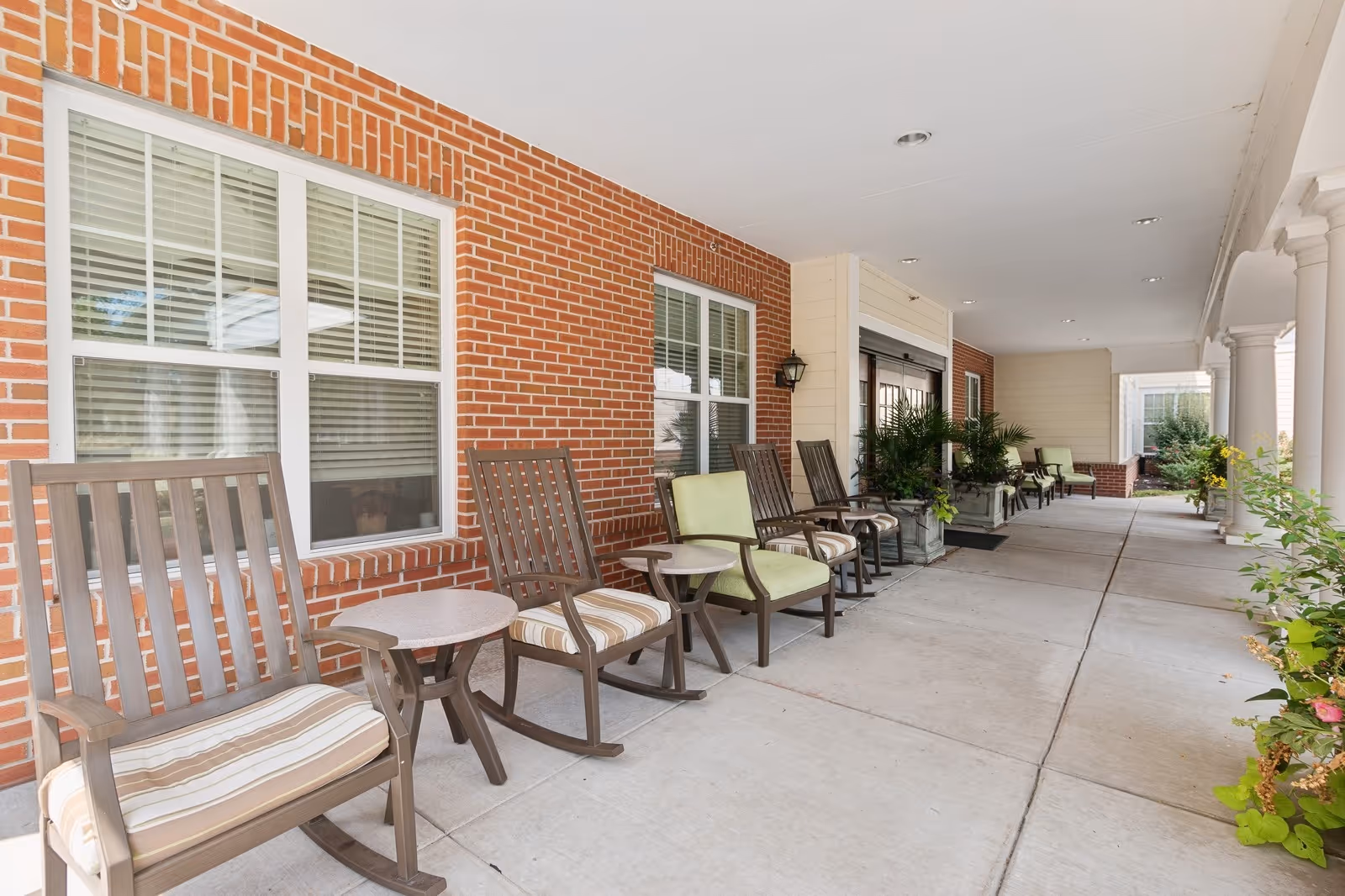 Covered outdoor patio area at The Bridges at Warwick with several wooden rocking chairs and small tables arranged along a brick wall with windows. There are potted plants and columns supporting the roof along the walkway.