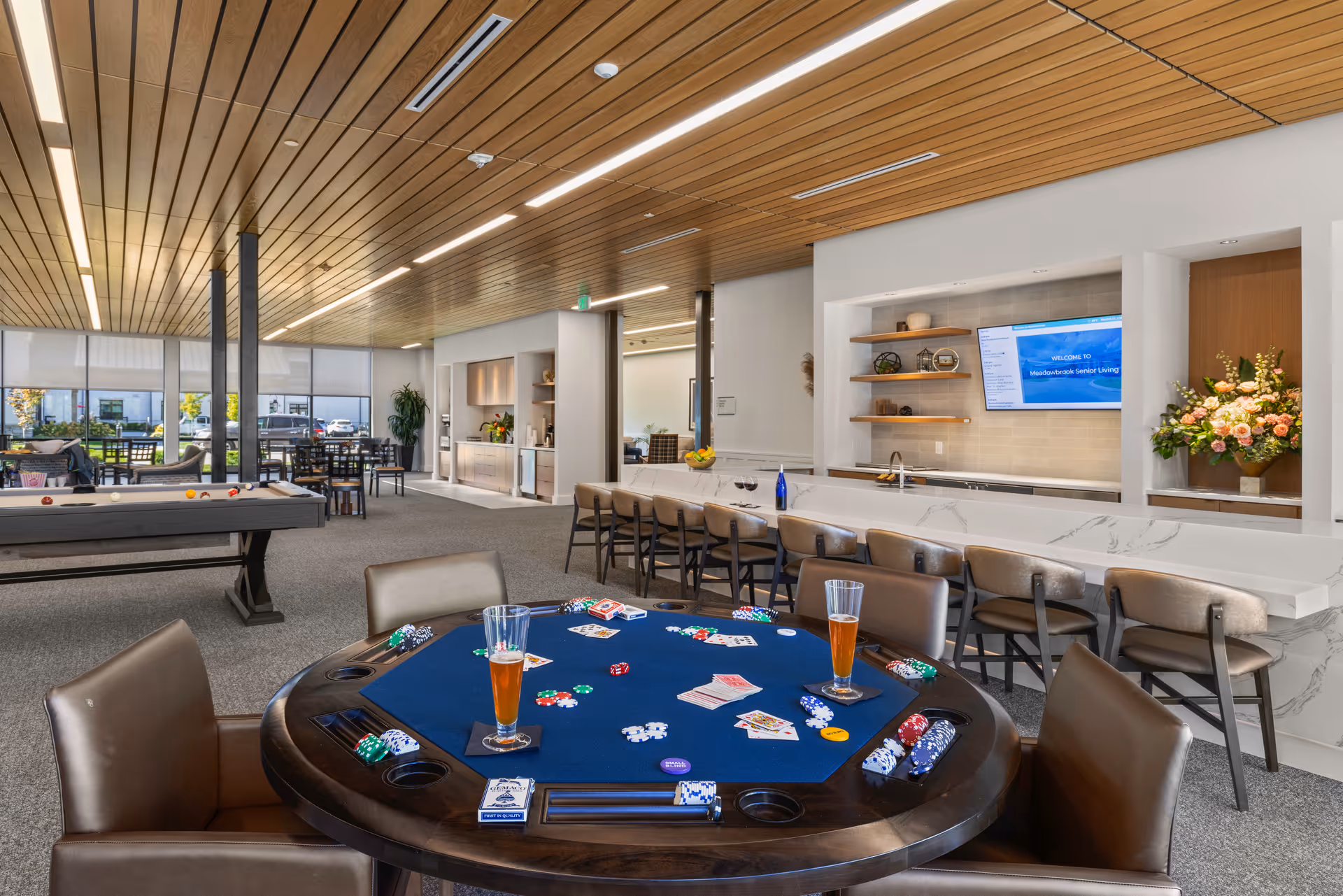 A spacious senior living common area featuring a poker table with playing cards, poker chips, and two glasses of beer. In the background, there is a pool table, several seating areas with tables and chairs, a long counter with bar stools, and a wall-mounted TV displaying a welcome message for Meadowbrook Senior Living. The room has a modern design with a wooden ceiling, large windows, and decorative plants and flowers.