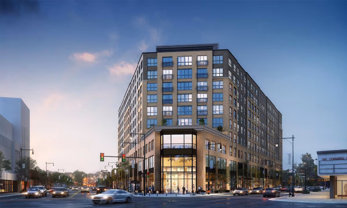 Large multi-story mixed-use building at an urban intersection with cars and pedestrians at dusk.