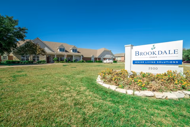 Exterior view of Brookdale Ennis senior living facility with a large green lawn, a clear blue sky, and a white sign in the foreground that reads 'Brookdale Ennis Senior Living Solutions 2500'. The building has a beige exterior with multiple windows and a sloped roof.