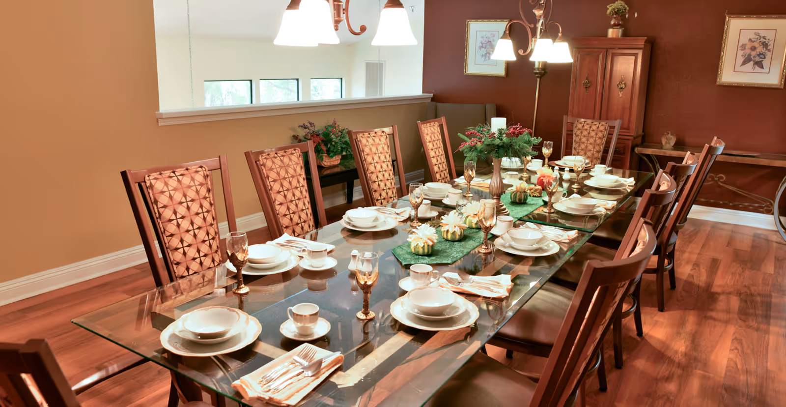 A long glass dining table set with dishes, glassware and centerpieces surrounded by upholstered chairs in a formal dining room.