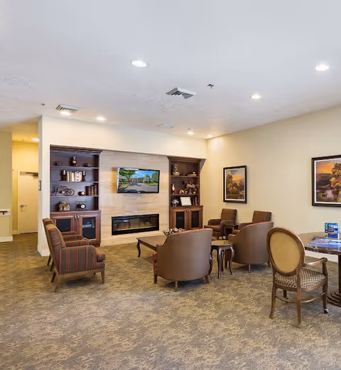 A cozy living room area in Meadow Creek Village Assisted Living featuring a carpeted floor, several upholstered chairs arranged around a wooden coffee table, built-in wooden shelves with decorative items, a wall-mounted flat-screen TV above a modern fireplace, and framed landscape paintings on the walls.