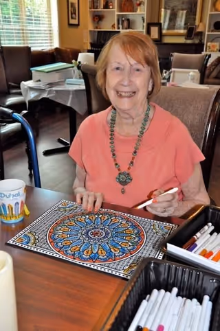 An elderly woman with short light brown hair wearing a coral shirt and a beaded necklace is sitting at a table in a well-lit room. She is smiling and coloring a detailed mandala design with markers. The table has a cup and a container filled with various colored markers. In the background, there are chairs, a window with blinds, and shelves with decorative items.