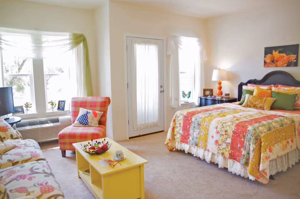 A bright and cozy bedroom with a colorful patchwork quilt on the bed, multiple pillows, a bedside table with a lamp, a red plaid armchair with a decorative pillow, a yellow coffee table with a cup and bowl, a floral patterned sofa, and large windows with sheer curtains letting in natural light.