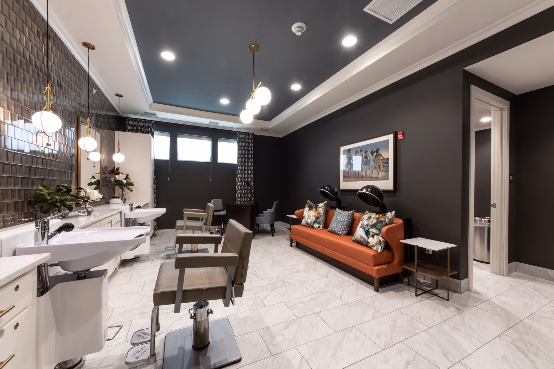 A modern salon area with two styling chairs in front of sinks and mirrors on the left side. The room has a dark ceiling with recessed lighting and pendant lights above the sinks. On the right side, there is an orange sofa with floral and patterned pillows, two black hair dryers, and a framed picture on the dark wall. The floor is covered with white marble tiles, and there is a small table next to the sofa. A doorway is visible on the right side of the room.