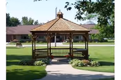 Wooden gazebo on a paved walkway in a grassy yard with a low building visible in the background.