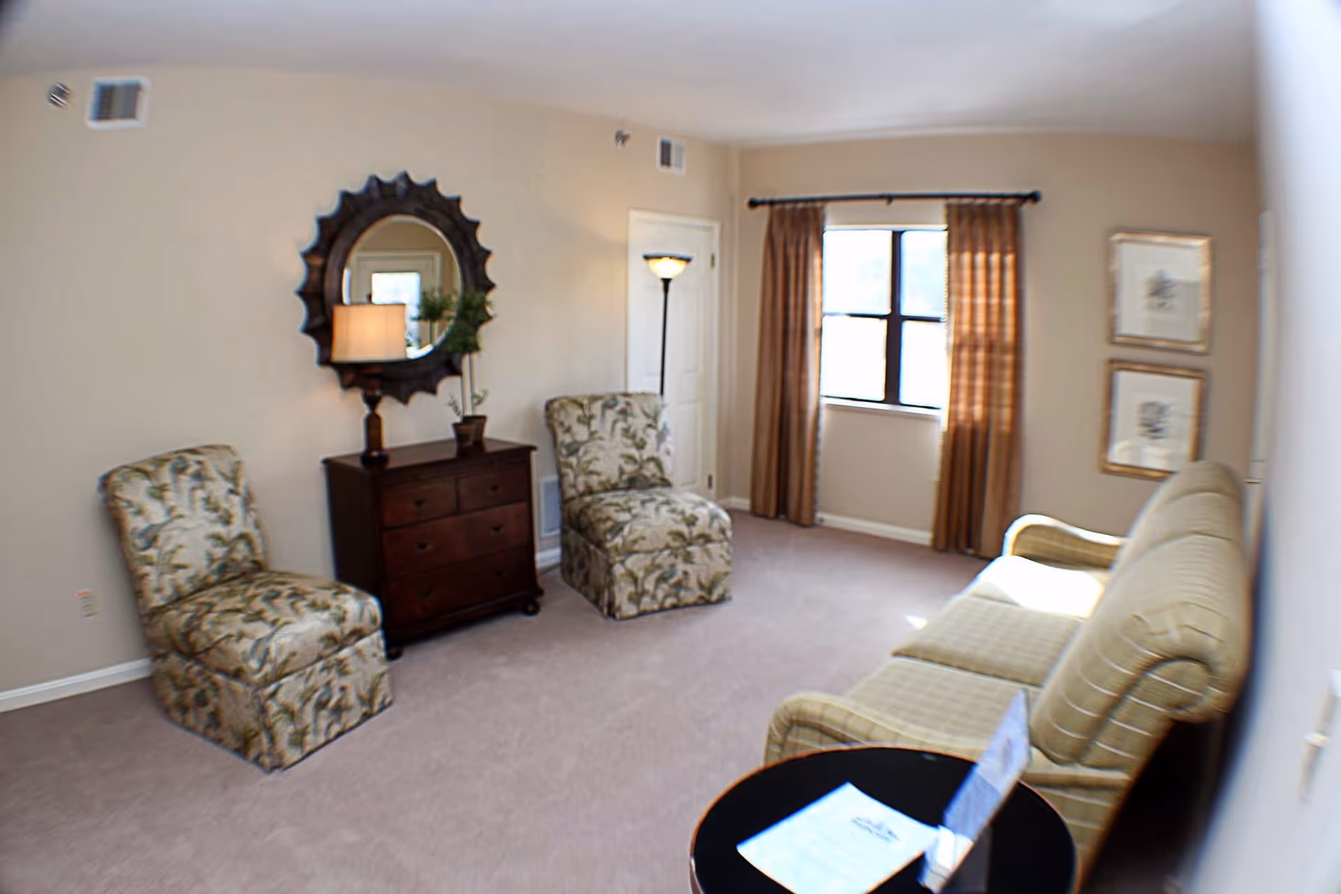 Bright furnished living room with patterned chairs, a sofa, a small dresser topped by a round mirror and lamp, and a curtained window.