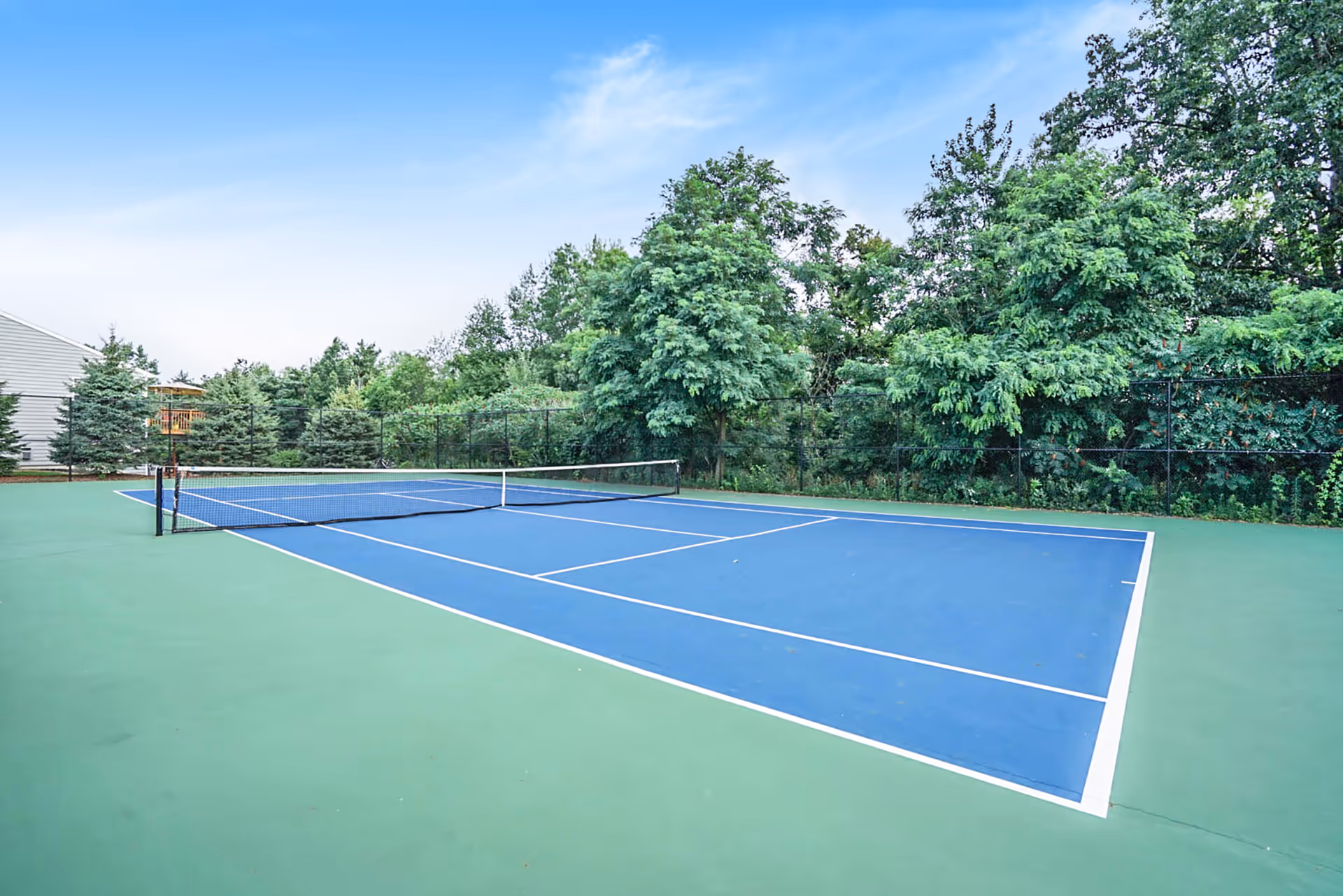 Outdoor tennis court with a blue playing surface and green surrounding area, enclosed by a black fence, with trees and a clear blue sky in the background.