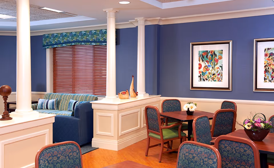 Interior view of a senior living facility common area with blue walls and wooden flooring. The room features several tables with floral-patterned chairs, decorative vases on a white half-wall with columns, a blue couch with striped pillows near a window with wooden blinds and a patterned valance, and colorful floral artwork on the walls.