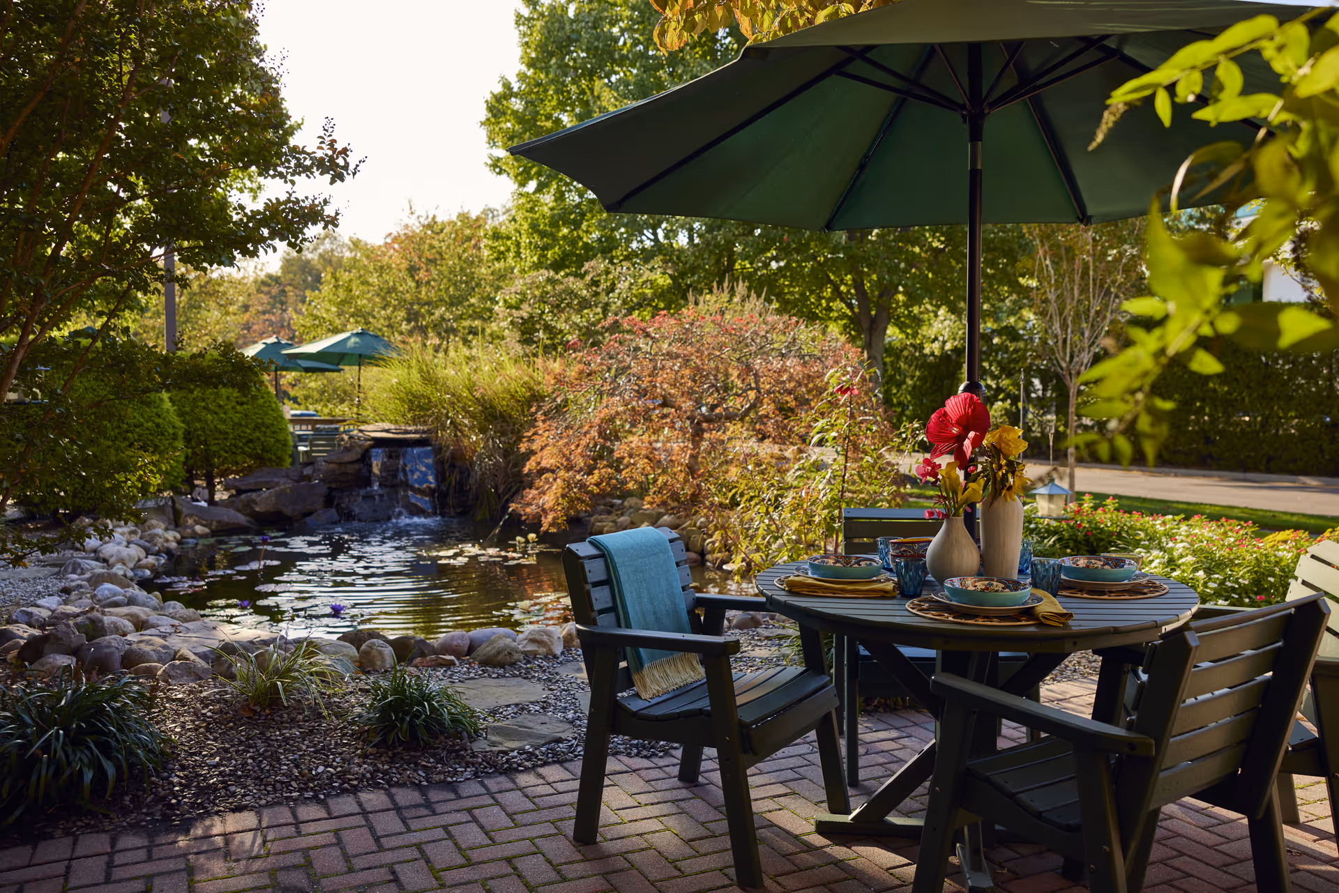 Outdoor patio area with a round table set for four, featuring colorful bowls, glasses, and vases with flowers. The table is shaded by a large green umbrella. Surrounding the patio are lush green trees, bushes, and a small pond with a waterfall and rocks, creating a serene garden setting.