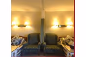 Two patterned armchairs placed side by side in a room with beige walls, each flanked by a hospital-style bed with floral pillows. Wall-mounted lights illuminate the area above the chairs.