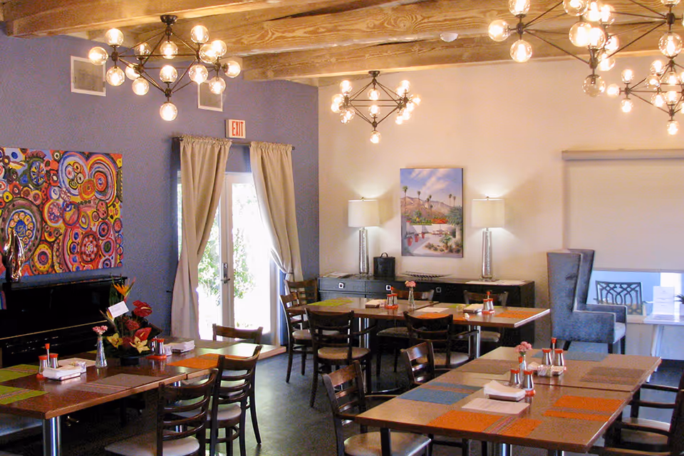 A dining room with multiple wooden tables and chairs, colorful placemats, and condiment sets on the tables. The room has modern chandeliers with exposed bulbs hanging from a wooden beam ceiling. There is a large colorful abstract painting on a purple wall, a door with curtains leading outside, and a sideboard with two lamps and a framed picture on the wall.