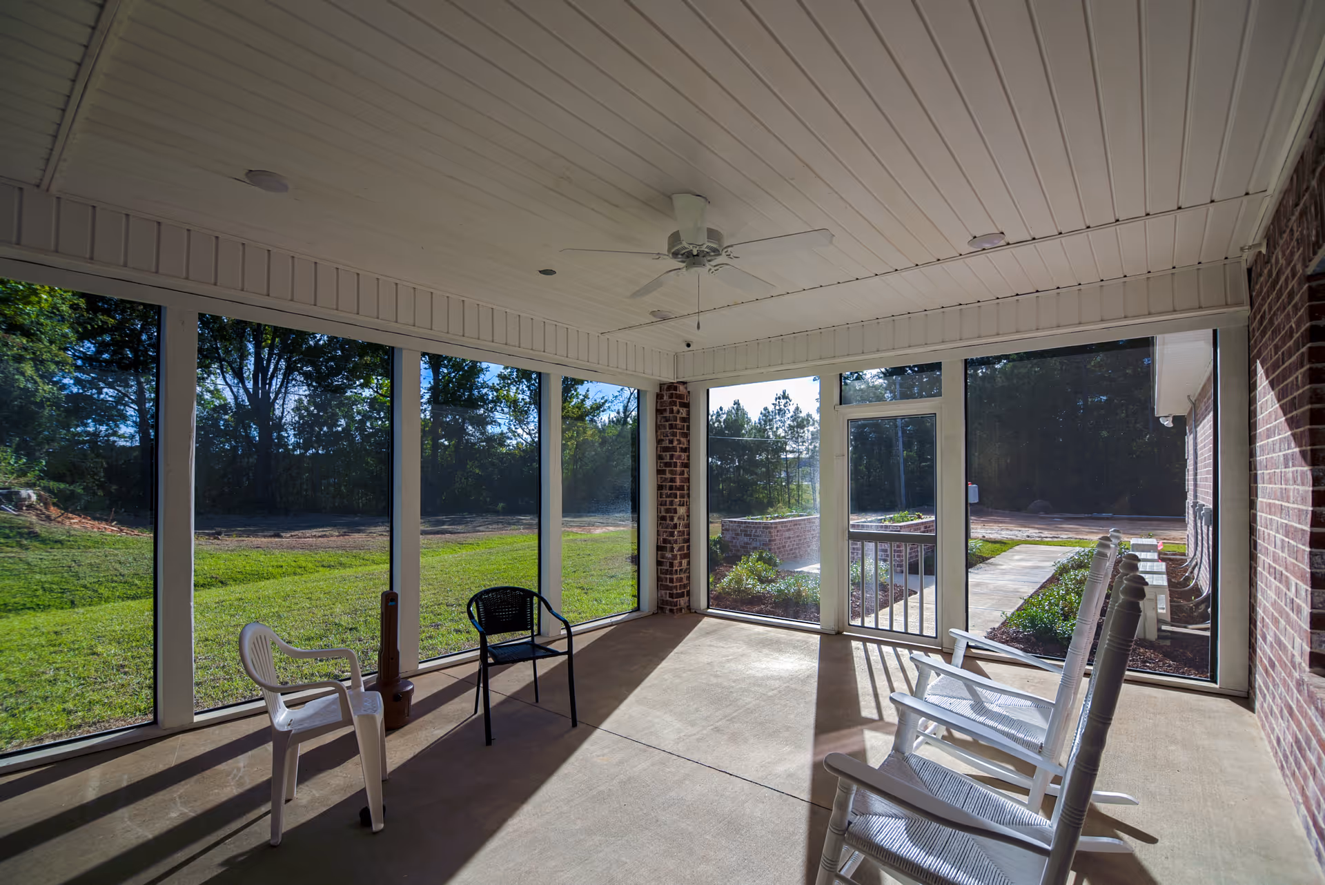 A screened-in porch area with a white ceiling fan, several chairs including white rocking chairs and plastic chairs, overlooking a grassy outdoor area with trees in the background.