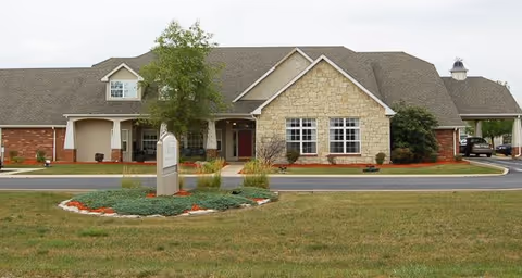 Front exterior view of a single-story senior living facility building with a combination of brick and stone facade, a covered entrance, and a landscaped circular driveway with a sign in the center.