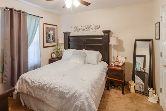 A cozy bedroom with a large bed featuring a dark wooden headboard and white bedding. There are two bedside tables with lamps, a standing mirror leaning against the wall, and decorative items including framed pictures and small statues. The room has beige walls, a ceiling fan with lights, and a window with brown and teal curtains.