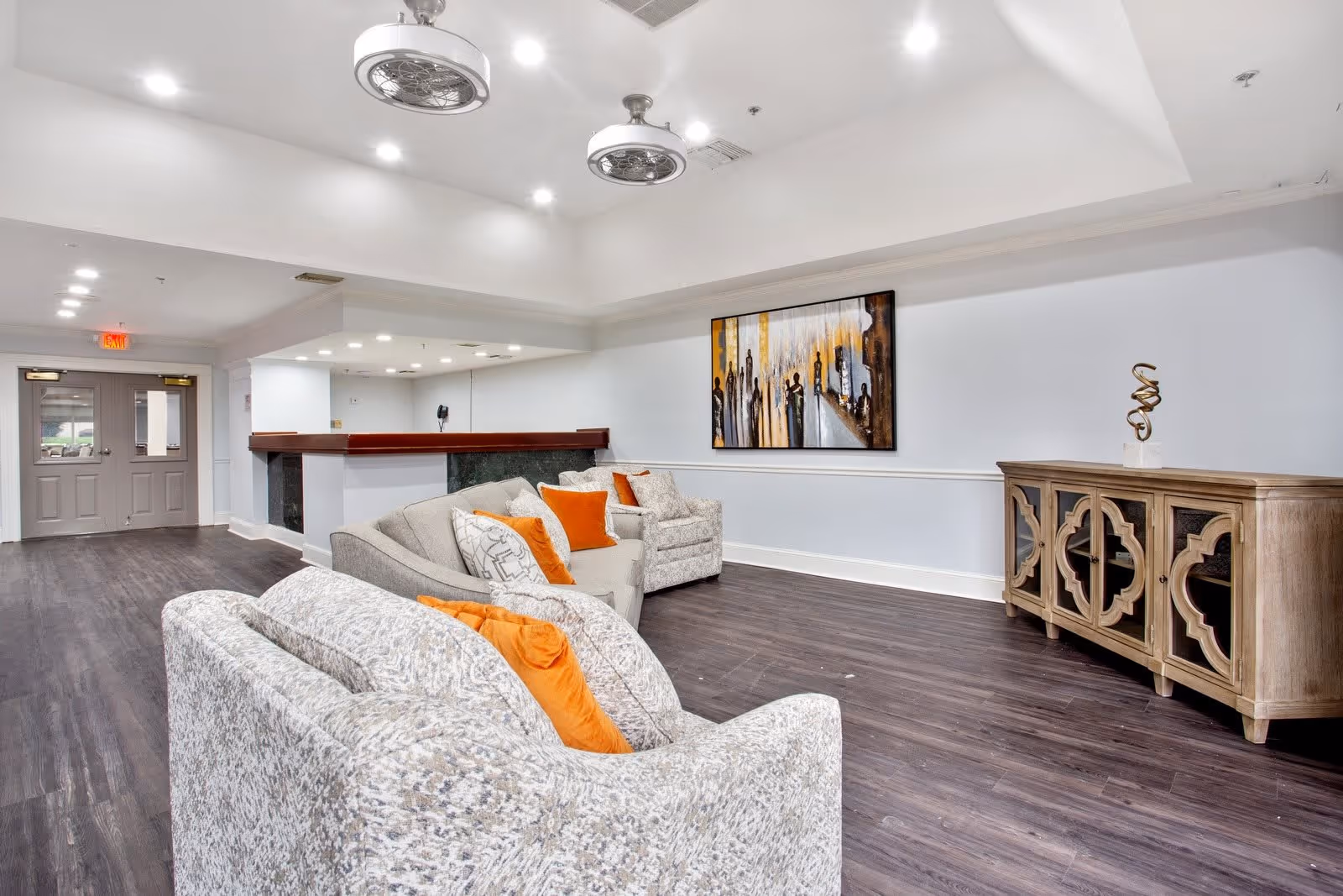 Bright communal lounge with upholstered sofas and orange pillows, a console table, wall art, and a reception counter.