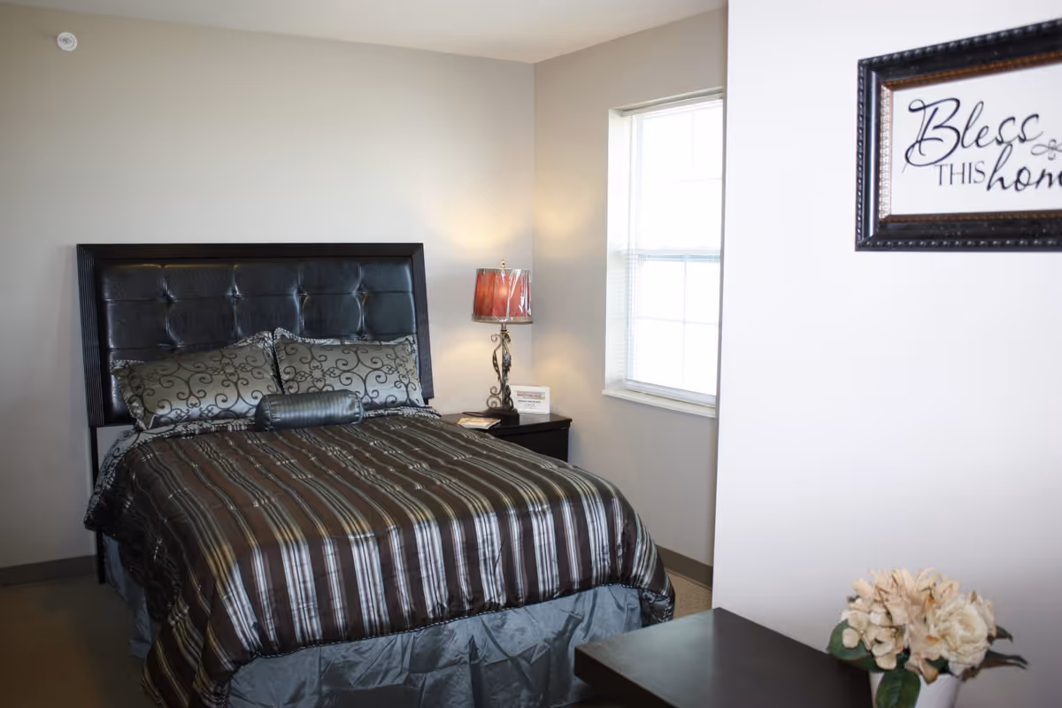 A bedroom with a bed featuring a dark padded headboard, striped brown and gray bedding, and decorative pillows. There is a nightstand with a lamp and a small card next to a window with blinds. A framed sign on the wall reads 'Bless this home,' and a vase with white flowers is on a nearby surface.