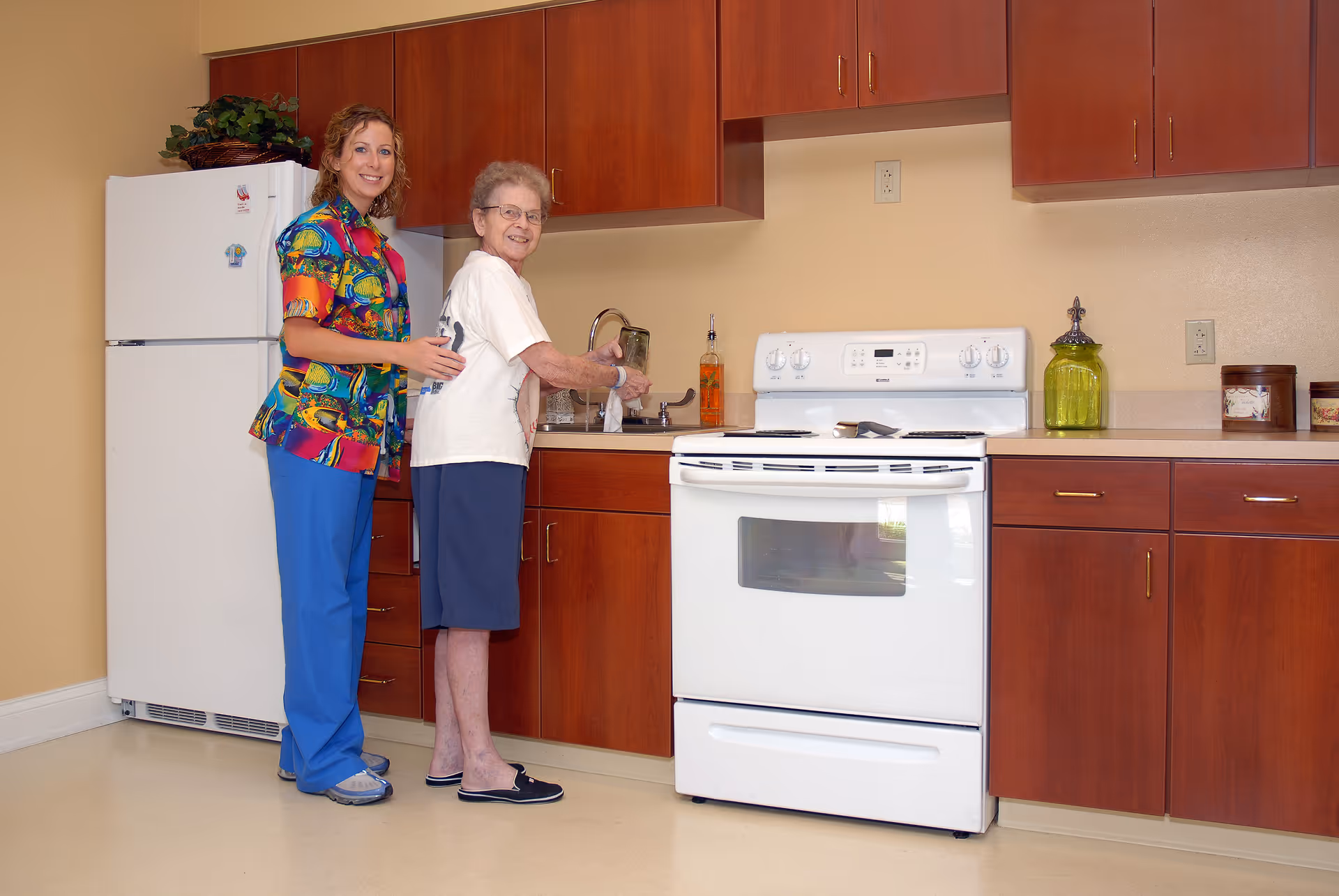 An elderly woman washing her hands at a kitchen sink with a caregiver standing beside her in a kitchen area with wooden cabinets, a white stove, and a white refrigerator.