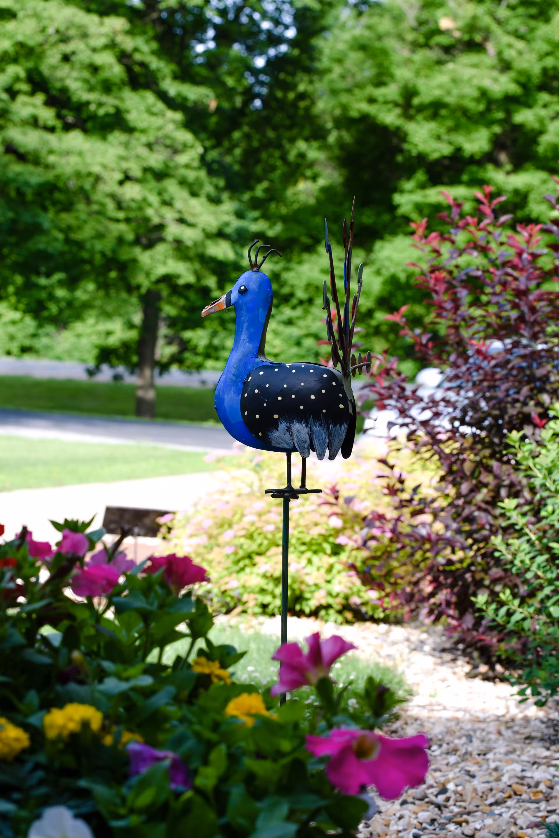 A colorful metal peacock garden ornament standing on a metal rod surrounded by vibrant flowers and greenery in an outdoor garden area.