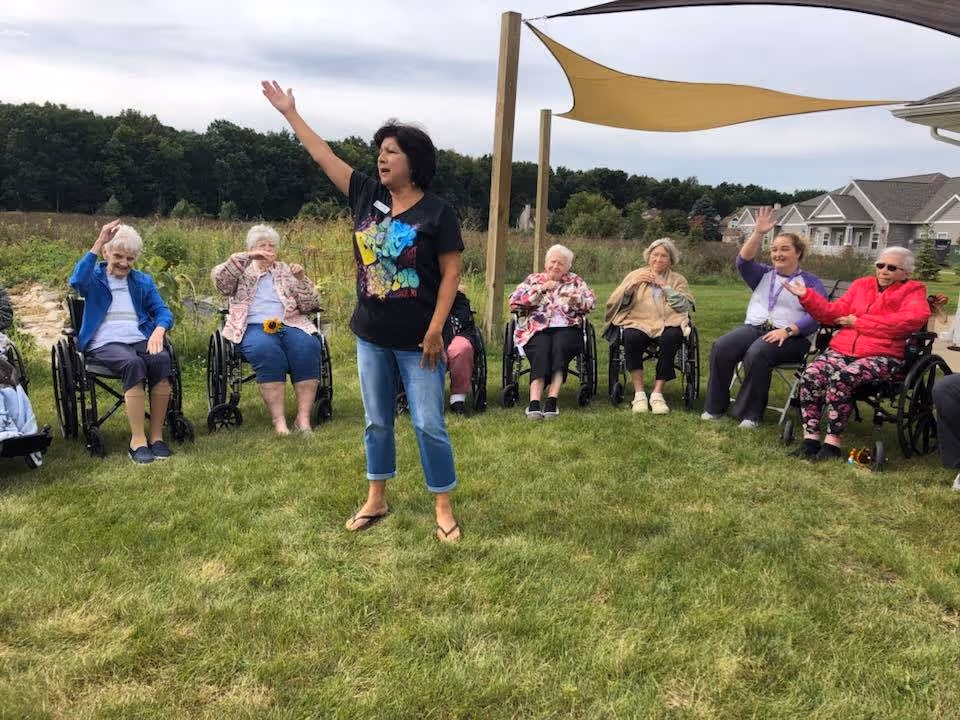 A group of elderly women, most in wheelchairs, sitting in a semi-circle on grass outdoors. A woman stands in the center with one arm raised, leading or engaging with the group. There are trees and houses in the background under a cloudy sky, with a shade sail overhead.