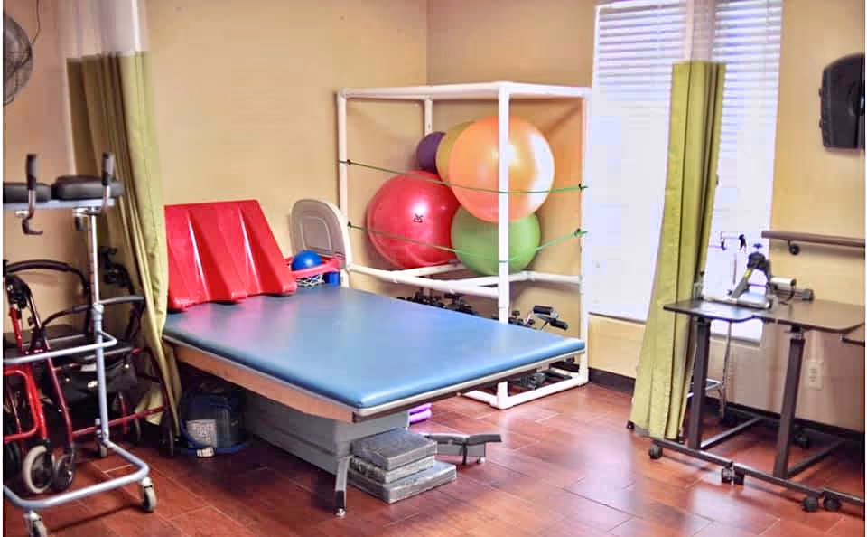 Therapy room with a treatment table, exercise balls on a rack, walkers and other mobility equipment near a window with green curtains.