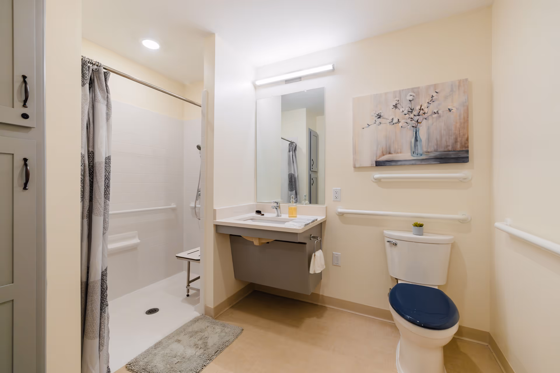 A clean and accessible bathroom with a walk-in shower featuring a shower chair and grab bars. The bathroom has a sink with a mirror above it, a toilet with a dark blue seat, and grab bars on the walls. There is a decorative painting of flowers in a vase hanging above the toilet.