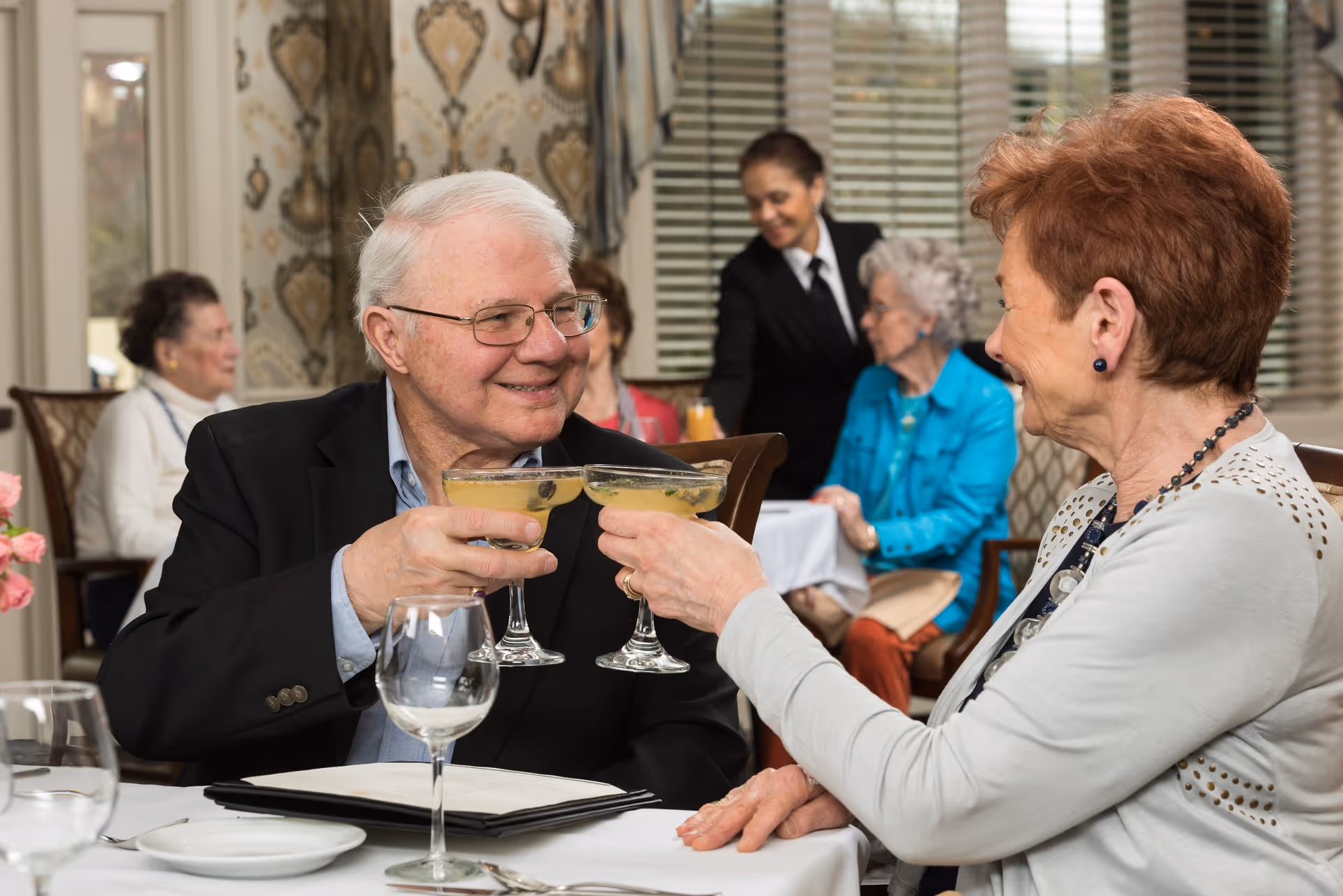 An elderly man and woman clink cocktail glasses and smile at each other while sitting at a dining table in a well-decorated room. In the background, other elderly people are seated and a server in a black uniform is attending to them.