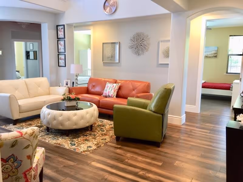 A cozy living room with a white sofa, a red leather sofa, a green armchair, and a floral patterned chair arranged around a round tufted ottoman with a glass top. The room has wooden flooring, a decorative rug, wall art, and a clock above the red sofa. Adjacent rooms with beds are visible through open doorways.