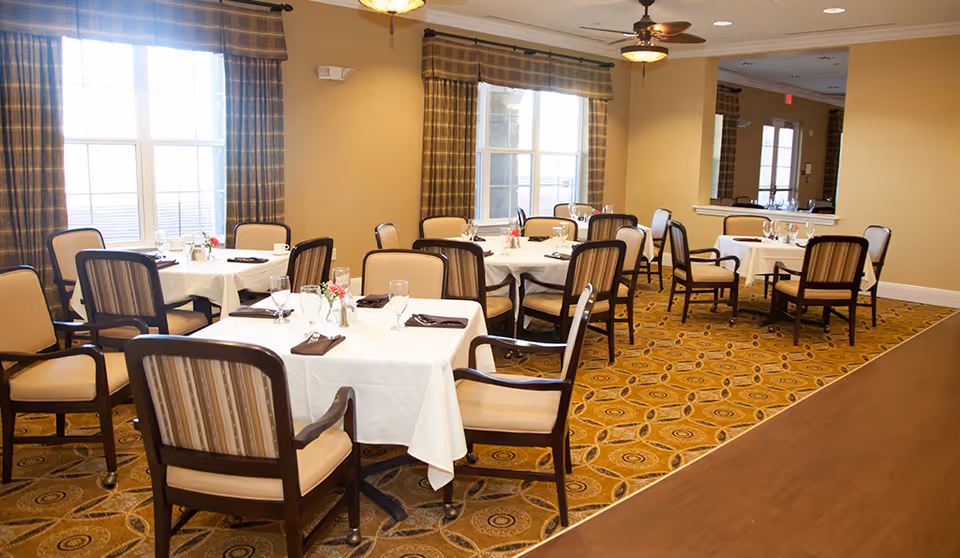 A bright dining room with several tables covered in white tablecloths, each set with glasses, napkins, and small flower arrangements. The room has large windows with plaid curtains, beige walls, patterned carpet, and ceiling fans with lights.