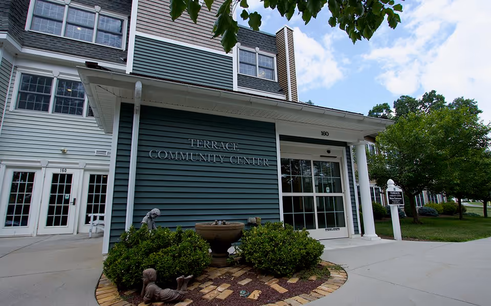 Exterior view of the Terrace Community Center building at Westminster Woods, featuring blue siding, white trim, a glass entrance door, and surrounding greenery including bushes and trees under a partly cloudy sky.