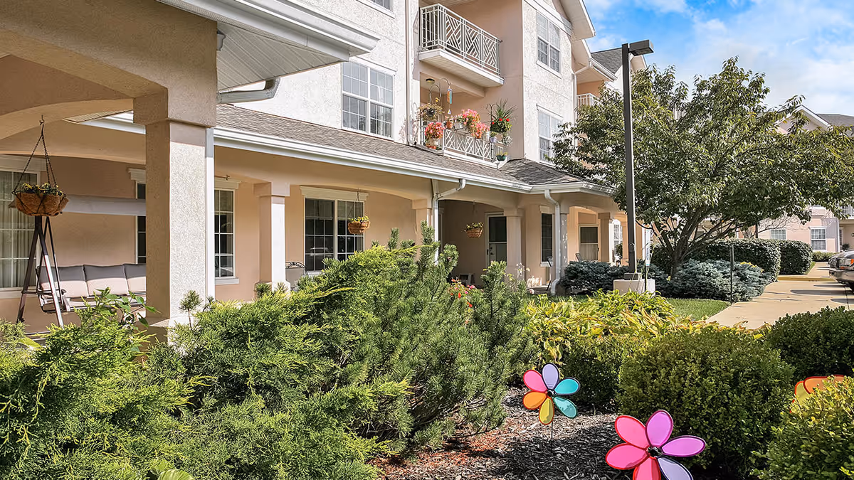 Exterior view of a senior living facility with beige walls and white trim. The building has balconies with flower pots and a covered porch area with hanging plants and outdoor seating. In the foreground, there are green bushes, colorful pinwheel garden decorations, and a tree. A sidewalk and parking area are visible on the right side under a partly cloudy sky.