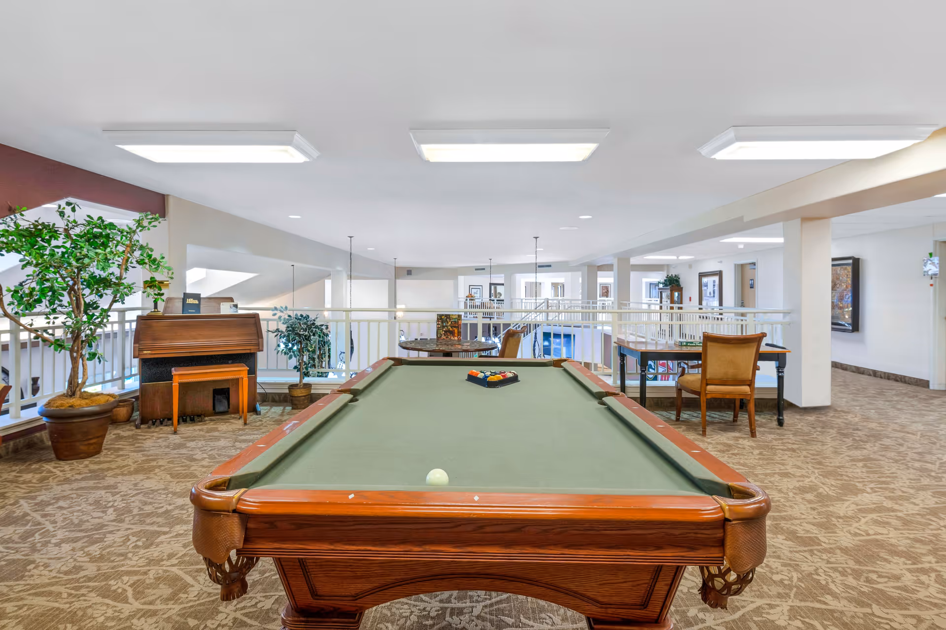 Interior view of a senior living facility's recreational area featuring a pool table with balls arranged for a game. The space has carpeted floors, a piano with a bench, potted plants, tables with chairs, and railings overlooking a lower level. The area is well-lit with ceiling lights and has a calm, inviting atmosphere.