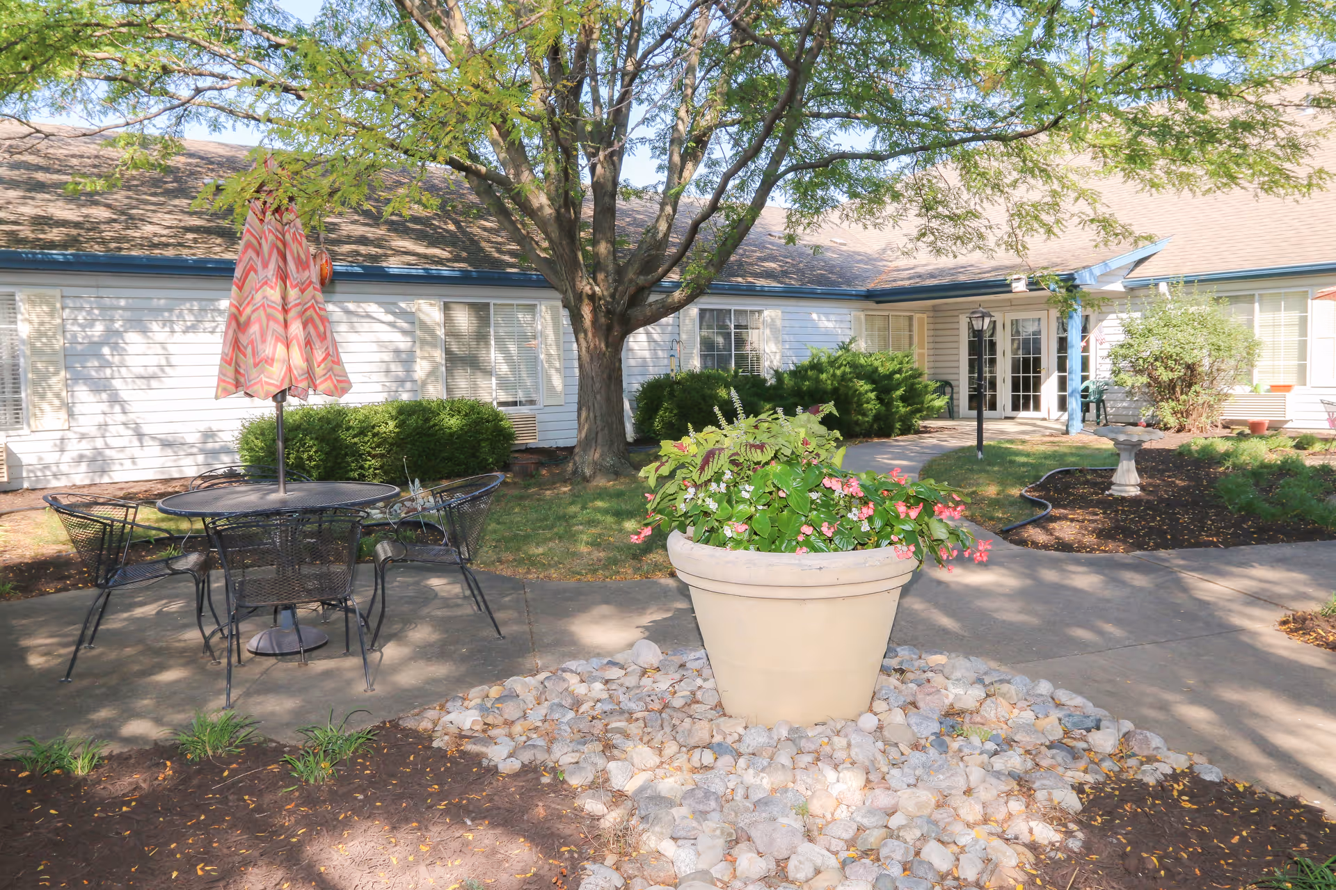 Outdoor patio area at Vintage Park at Lenexa featuring a round metal table with four matching chairs and a closed umbrella with a pink and white zigzag pattern. The patio is surrounded by greenery including bushes, a large tree, and a large planter with colorful flowers. The building exterior with white siding and multiple windows is visible in the background.
