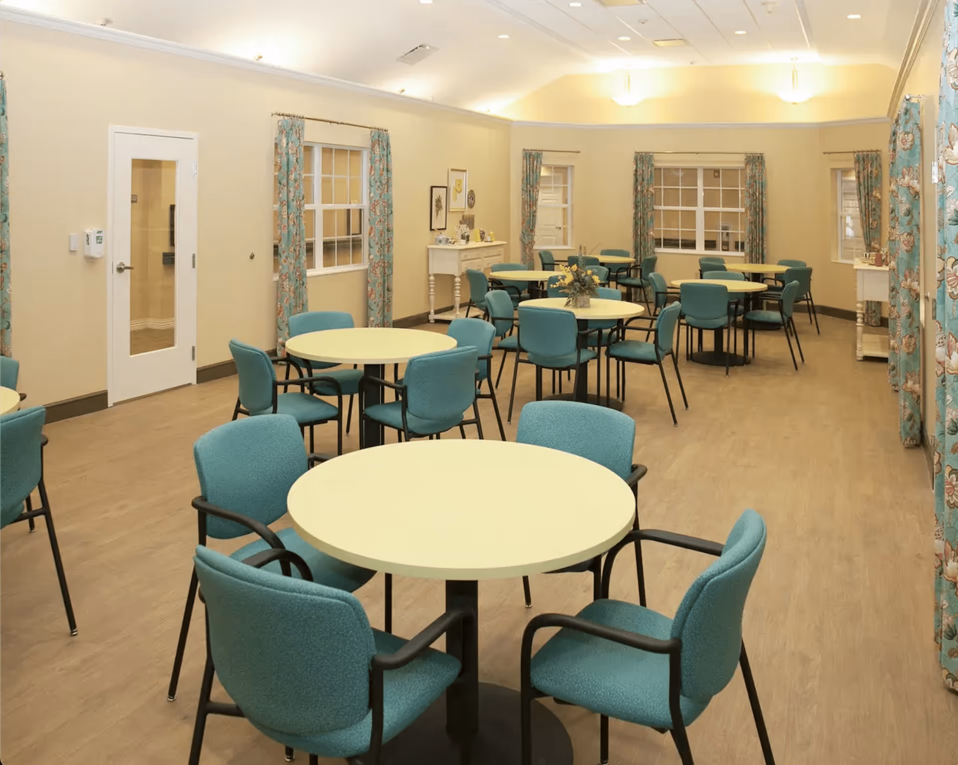 A spacious dining room with multiple round tables surrounded by teal upholstered chairs. The room has light yellow walls, wood flooring, and windows with floral curtains. There are decorative items on small tables along the walls.